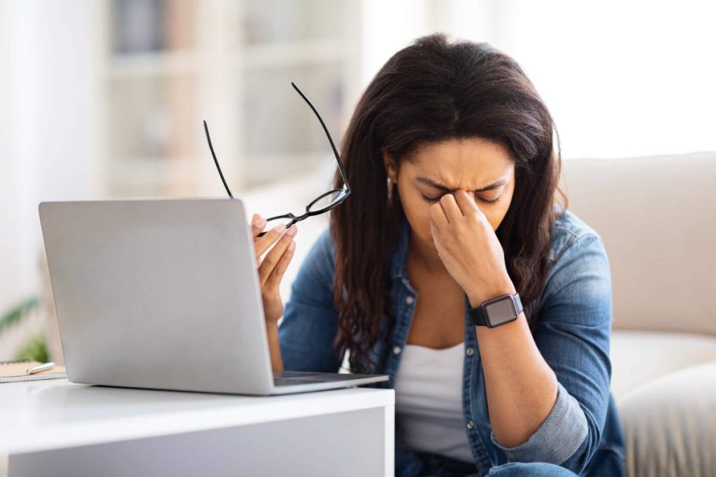 The image captures a black lady at home evidently stressed or overwhelmed, holding her glasses away from a laptop, illustrating common work-from-home challenges