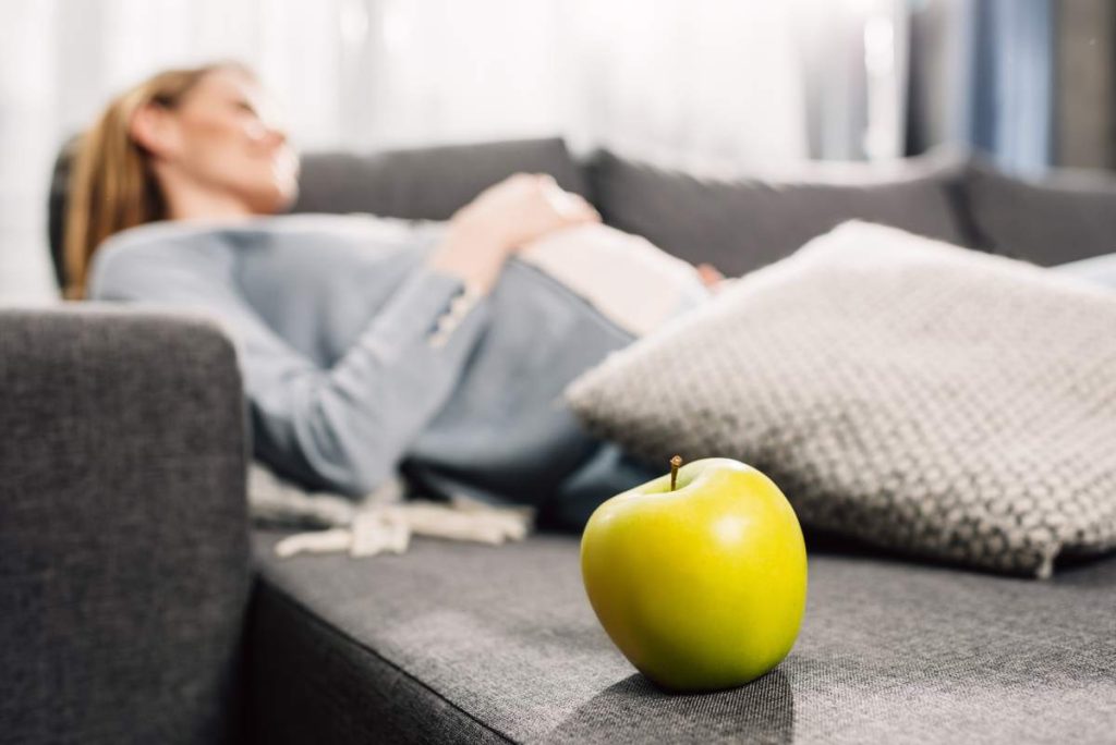 Young pregnant woman resting on gray sofa, green apple on table in foreground