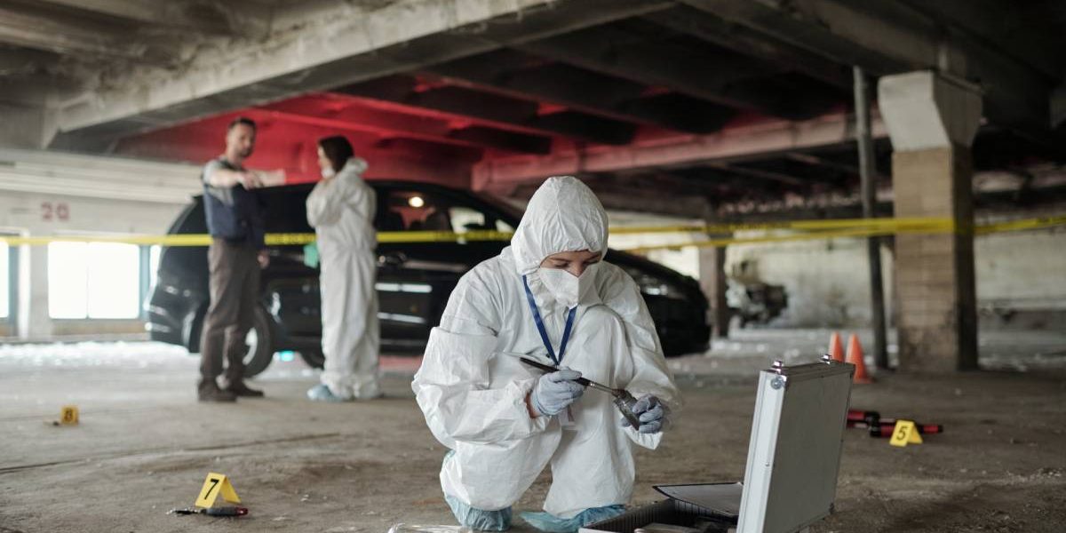 Young female criminological expert in protective workwear looking for fingerprints of murderer on crime scene against two colleagues