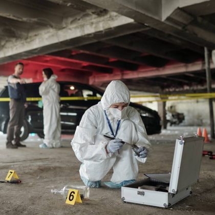 Young female criminological expert in protective workwear looking for fingerprints of murderer on crime scene against two colleagues