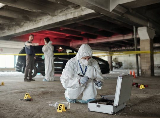 Young female criminological expert in protective workwear looking for fingerprints of murderer on crime scene against two colleagues