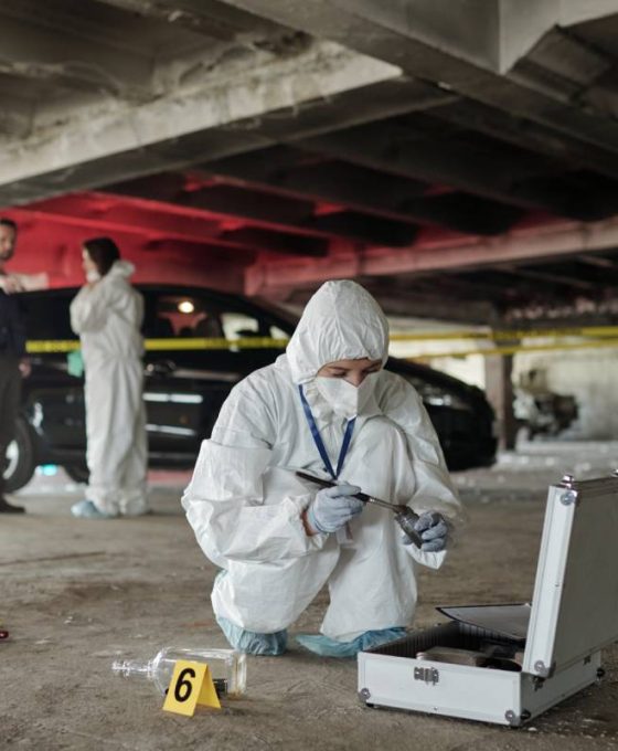 Young female criminological expert in protective workwear looking for fingerprints of murderer on crime scene against two colleagues