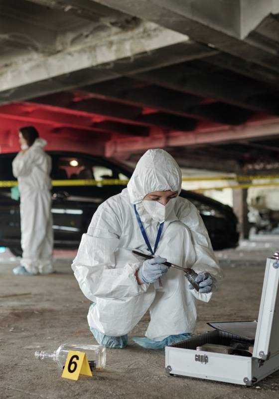 Young female criminological expert in protective workwear looking for fingerprints of murderer on crime scene against two colleagues