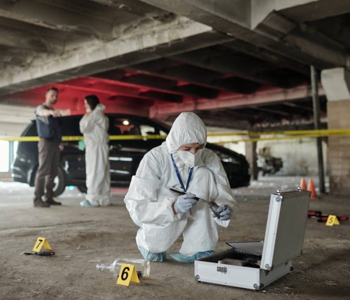 Young female criminological expert in protective workwear looking for fingerprints of murderer on crime scene against two colleagues