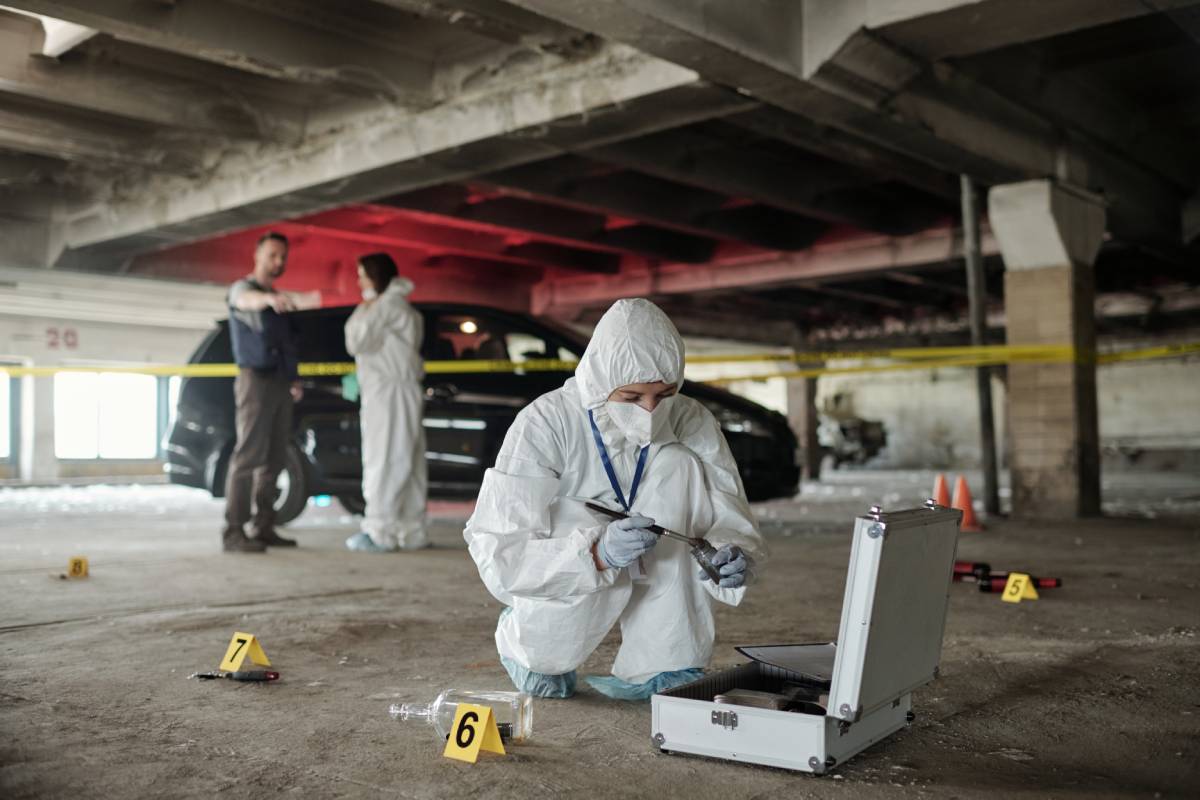 Young female criminological expert in protective workwear looking for fingerprints of murderer on crime scene against two colleagues